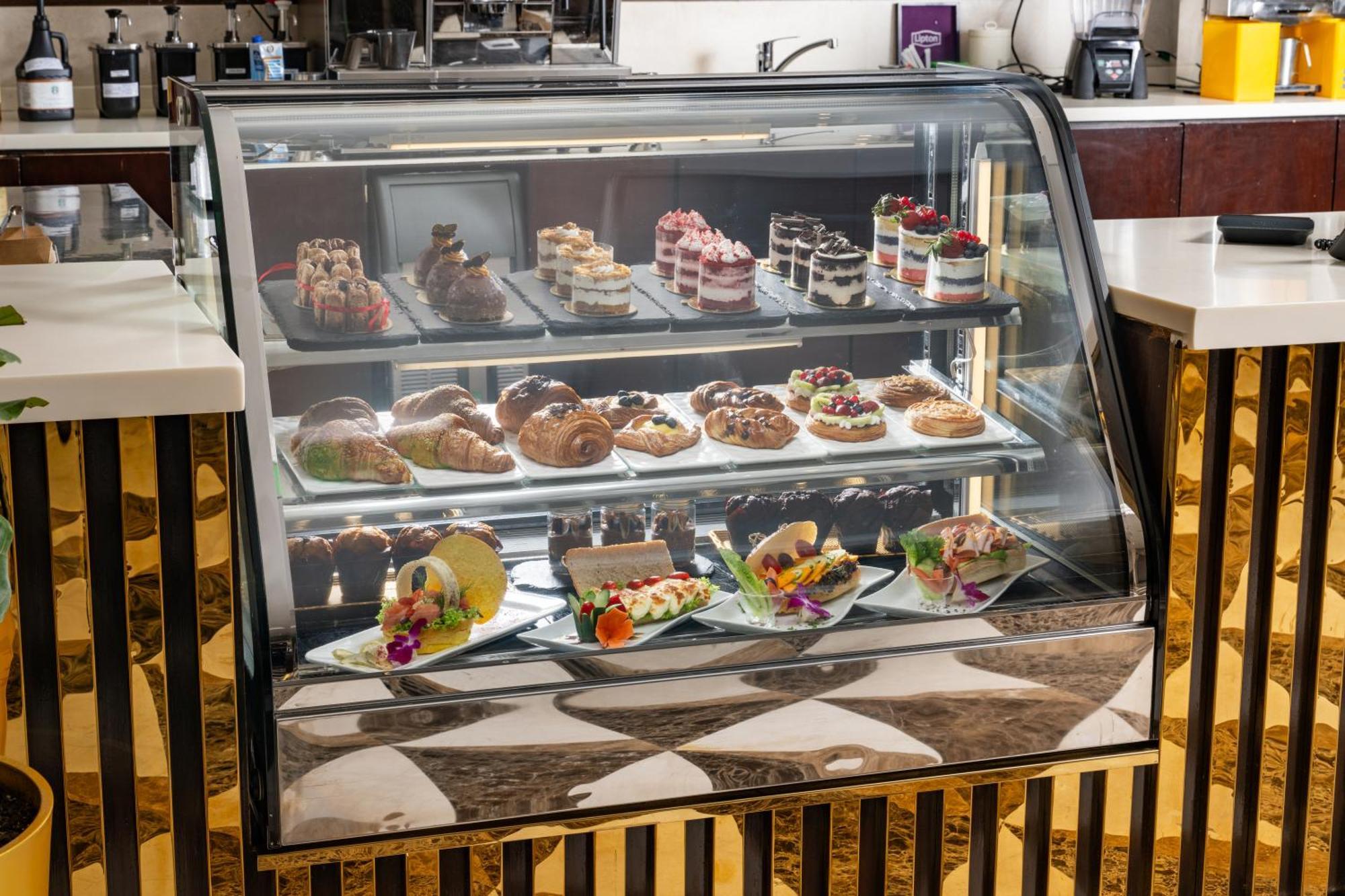 Al Aqeeq Madinah Hotel Медина Екстериор снимка The photo shows a display case filled with an assortment of desserts. There are various types of pastries, including cakes, tarts, and croissants, arranged neatly on different shelves. The desserts are visually appealing, with colorful decorations, f