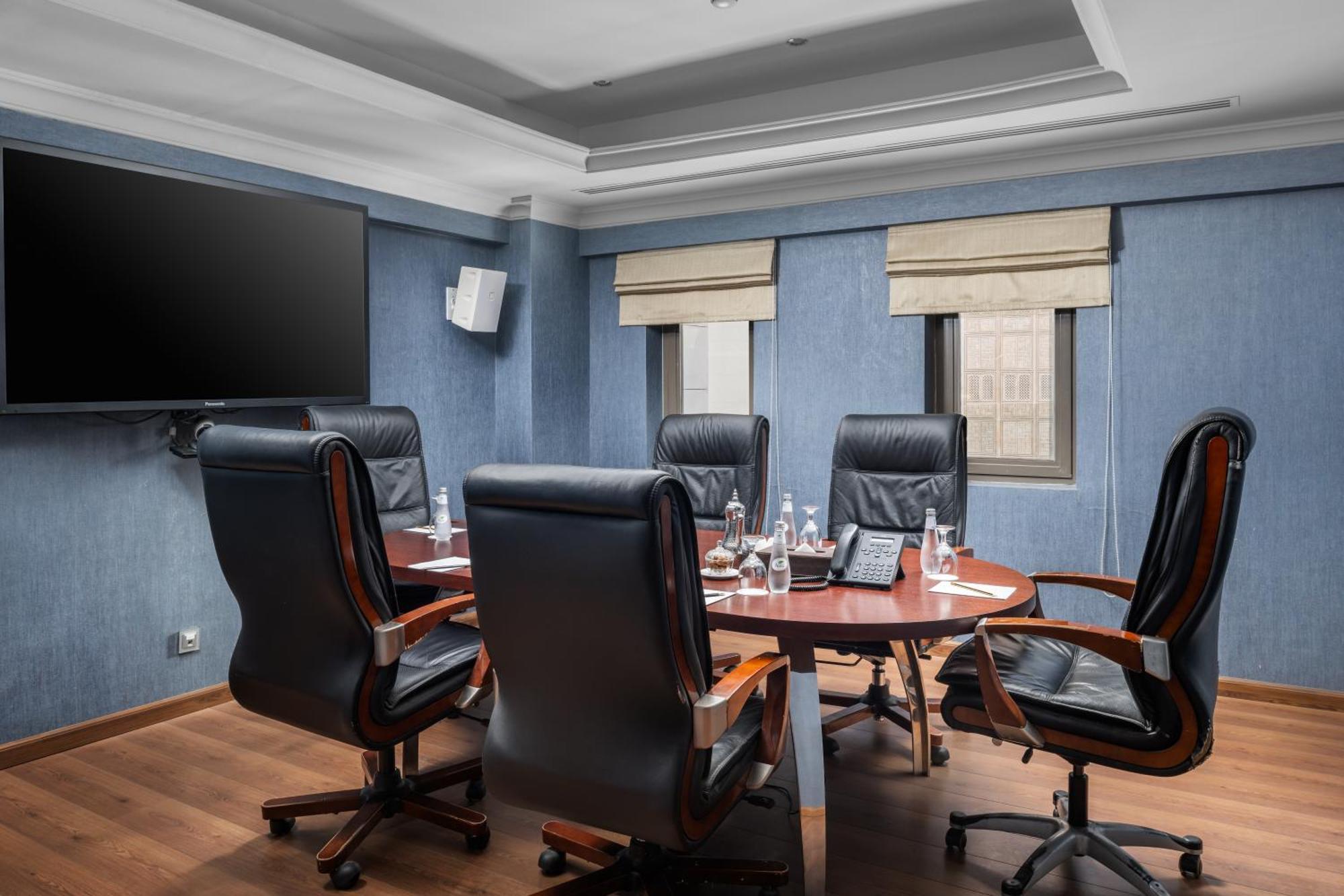 Al Aqeeq Madinah Hotel Медина Екстериор снимка The photo shows a modern conference room. It features a round wooden table surrounded by several black leather swivel chairs. The walls have a blue color scheme, and there is a large flat-screen TV mounted on one wall. The room has two windows with b