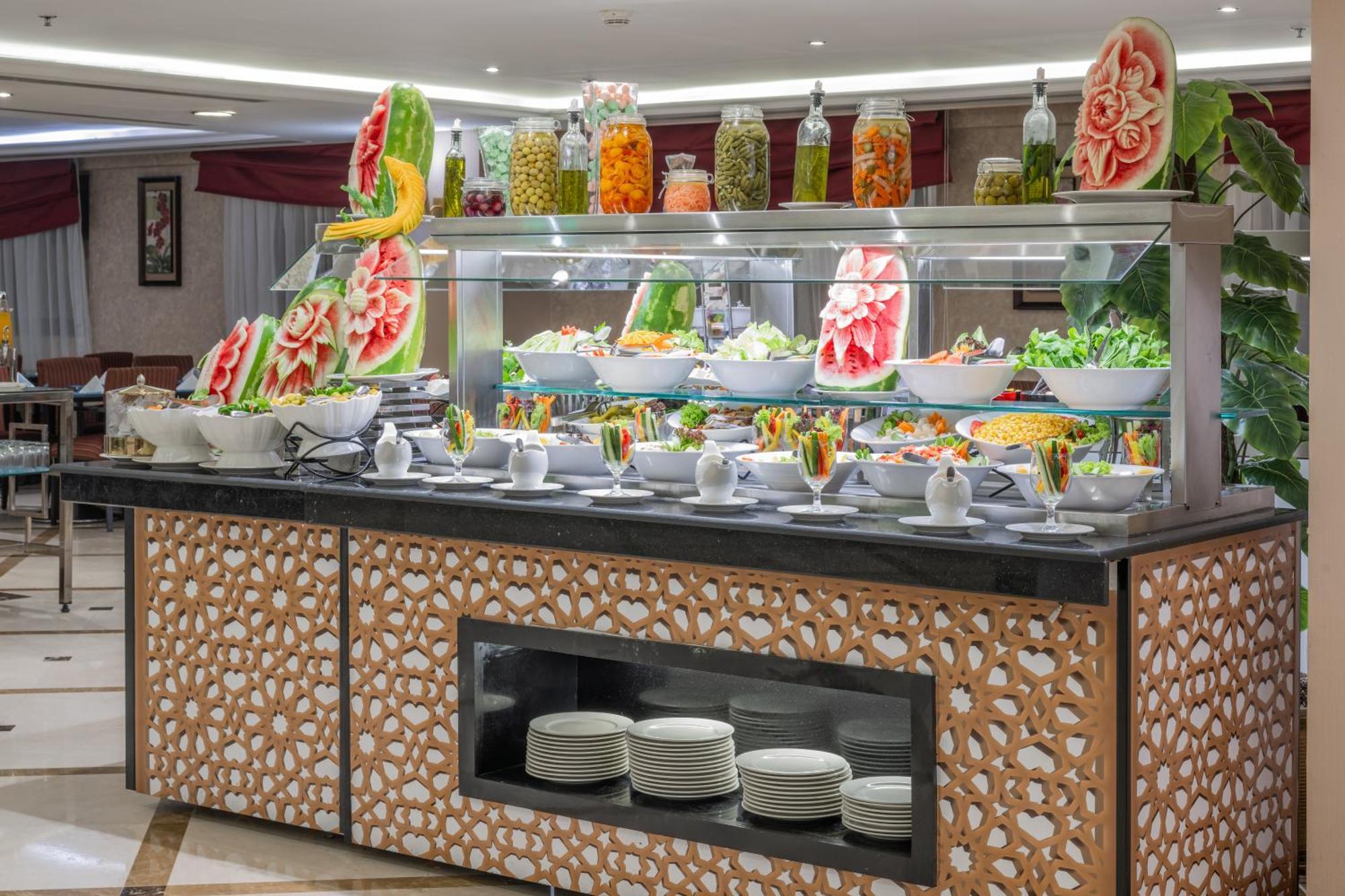 Al Aqeeq Madinah Hotel Медина Екстериор снимка The photo shows a buffet display that features an array of colorful food items. In the foreground, there are several large bowls filled with various salads and dishes. The presentation is visually appealing, with decorative watermelon carvings and an