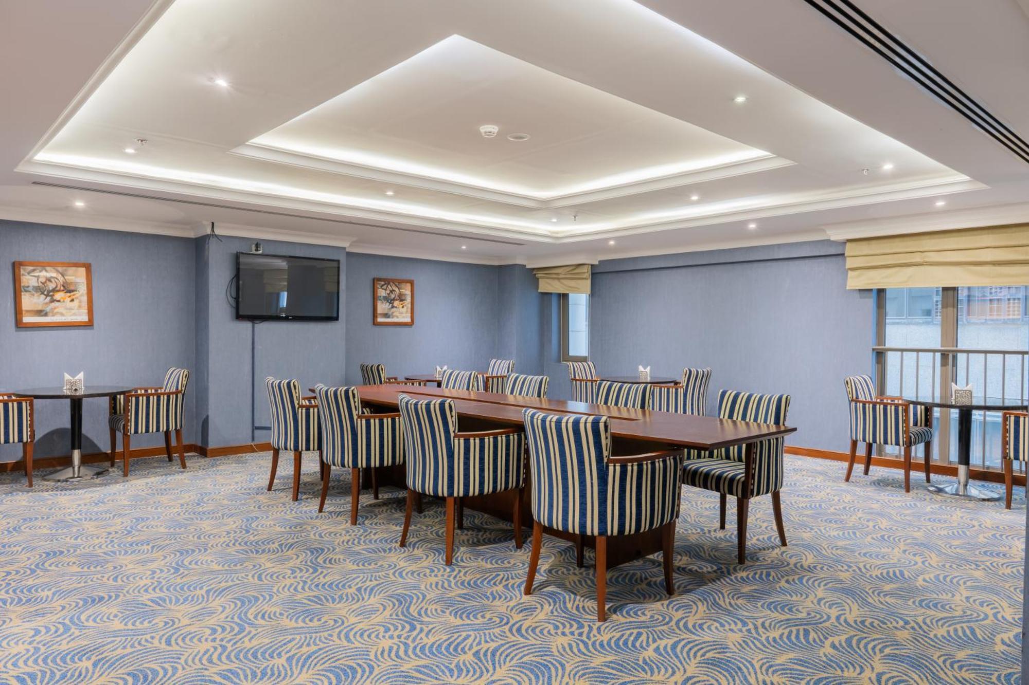 Al Aqeeq Madinah Hotel Медина Екстериор снимка The photo shows a spacious conference or meeting room. It features a light blue color scheme with a patterned carpet. There are several rectangular tables arranged in a group, surrounded by striped chairs. The room is well-lit with modern ceiling lig