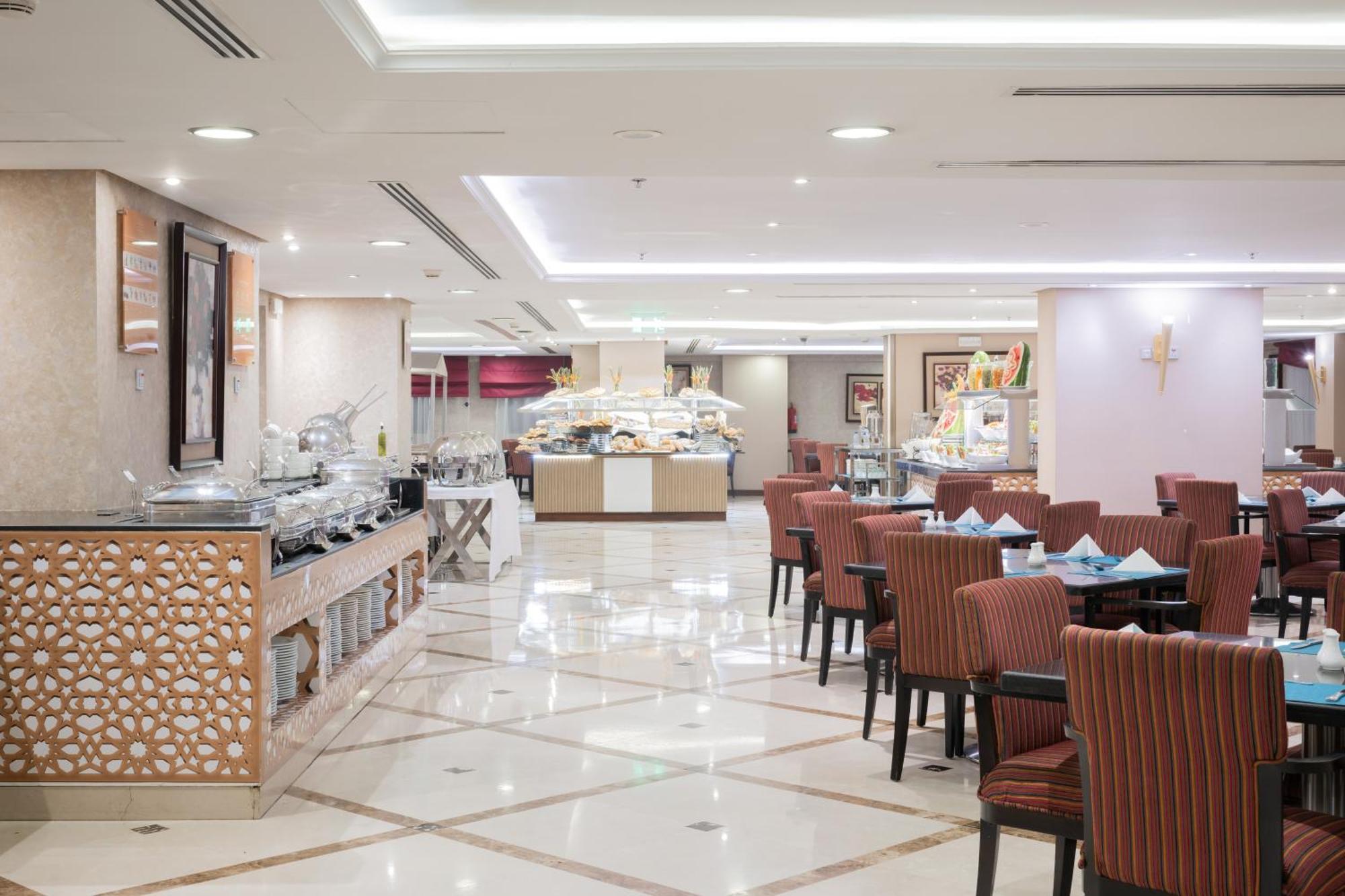 Al Aqeeq Madinah Hotel Медина Екстериор снимка The photo shows an elegantly designed restaurant or dining area. It features a spacious layout with a glossy tiled floor and bright, soft lighting. There are several dining tables with striped chairs arranged neatly. Along one side, there's a buffet 