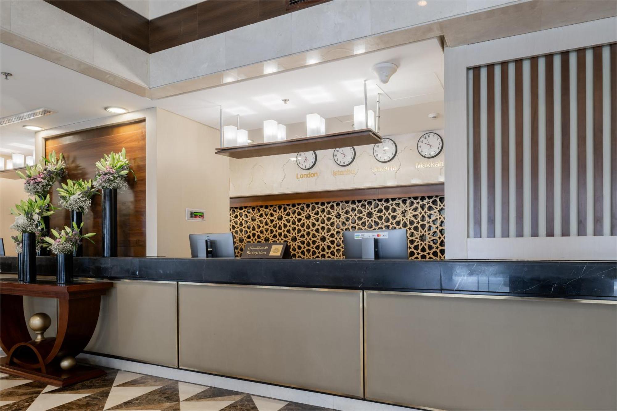 Al Aqeeq Madinah Hotel Медина Екстериор снимка The photo shows the interior of a hotel reception area. The reception desk is designed with a sleek, modern look featuring a dark countertop and a light-colored front. Above the desk, there are several clocks displaying different times, suggesting an