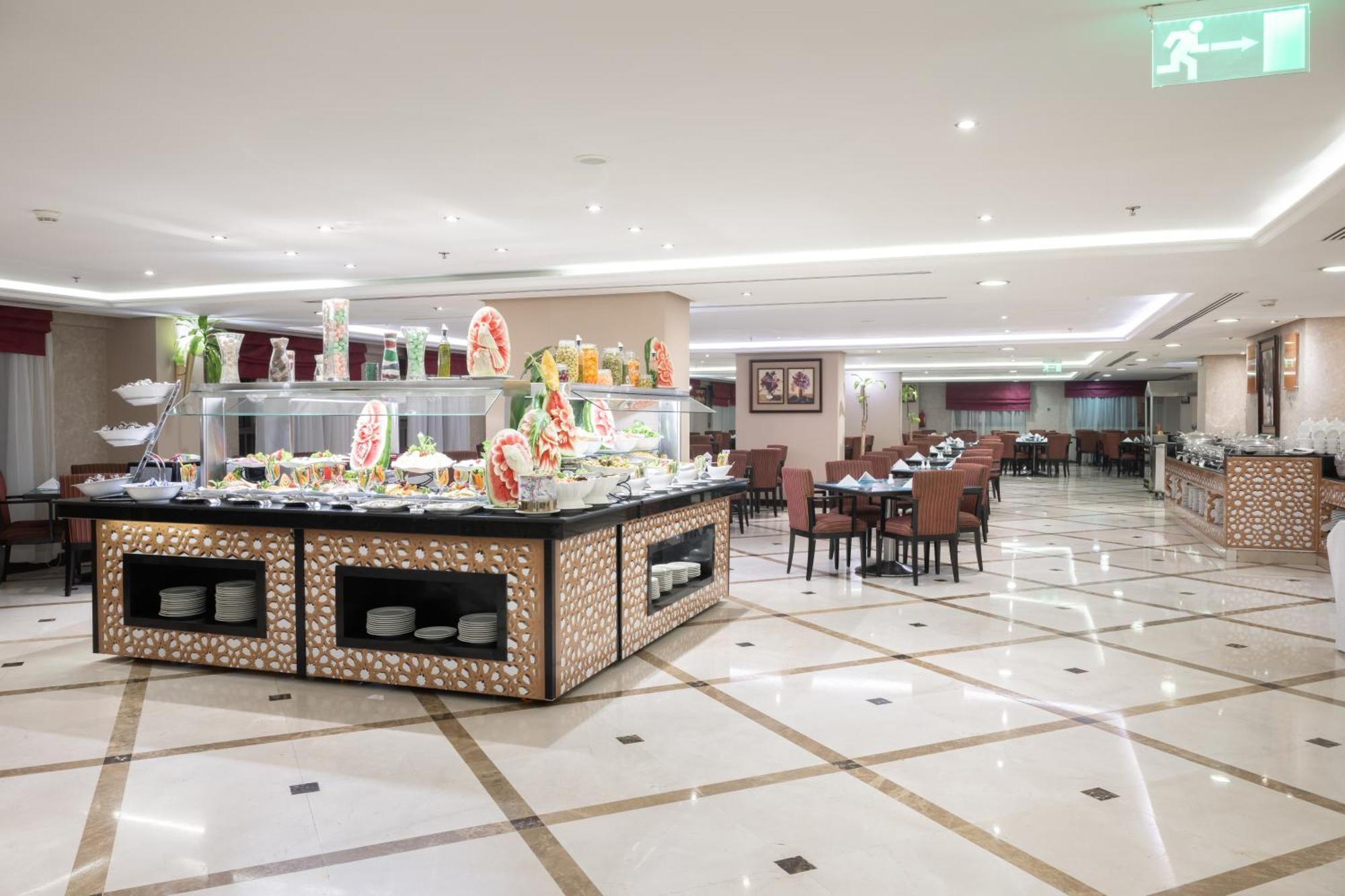 Al Aqeeq Madinah Hotel Медина Екстериор снимка The photo shows a spacious restaurant interior with a modern design. In the foreground, there is a large buffet counter filled with various food items, beautifully arranged and decorated. The counter features a backlit display, showcasing fruits and 