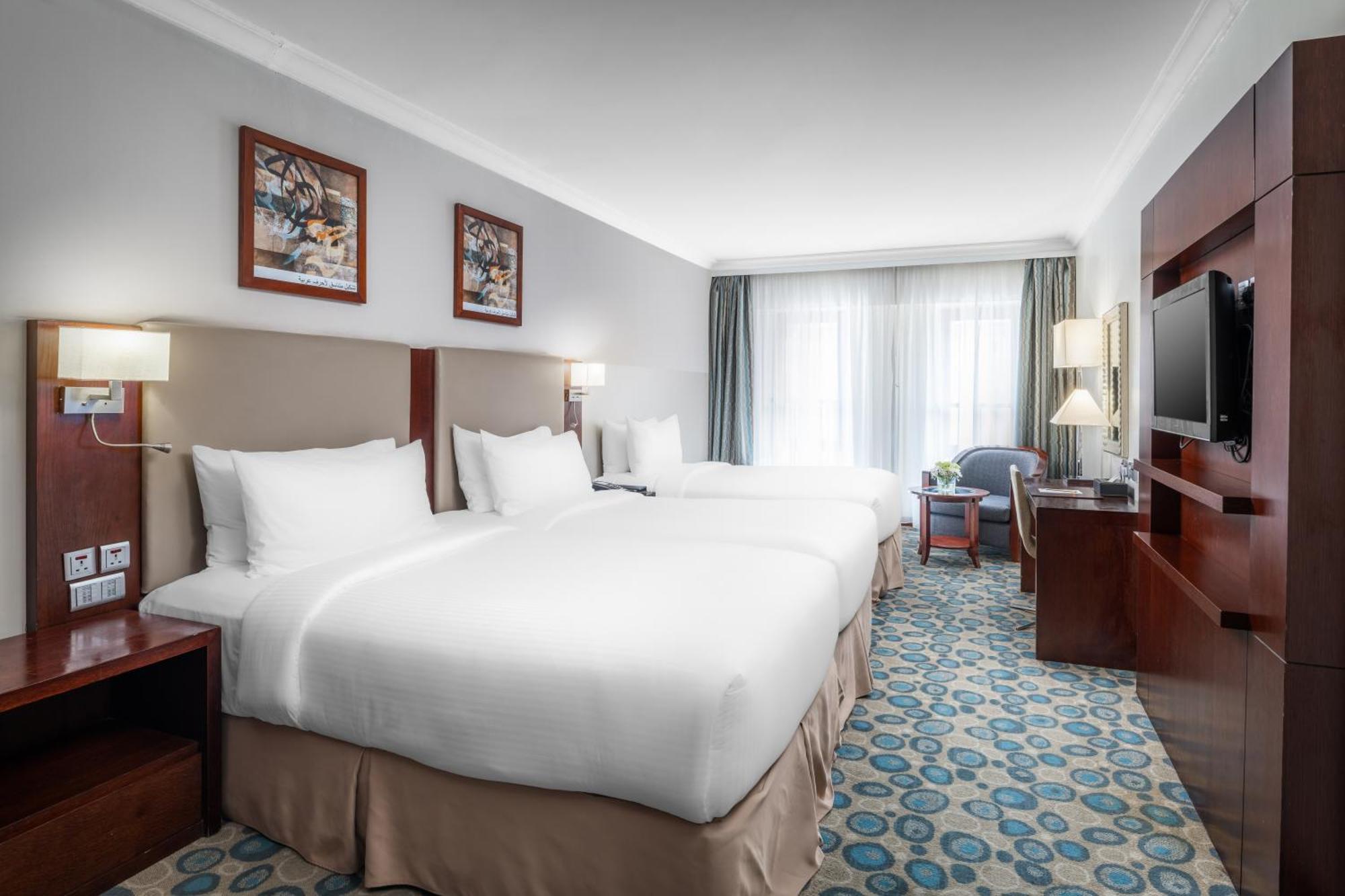 Al Aqeeq Madinah Hotel Медина Екстериор снимка The photo shows a well-furnished hotel room featuring two large beds with white bedding. The walls are adorned with framed artwork. There are bedside lamps next to each bed, and a flat-screen TV is mounted on the wall. A large window allows natural l