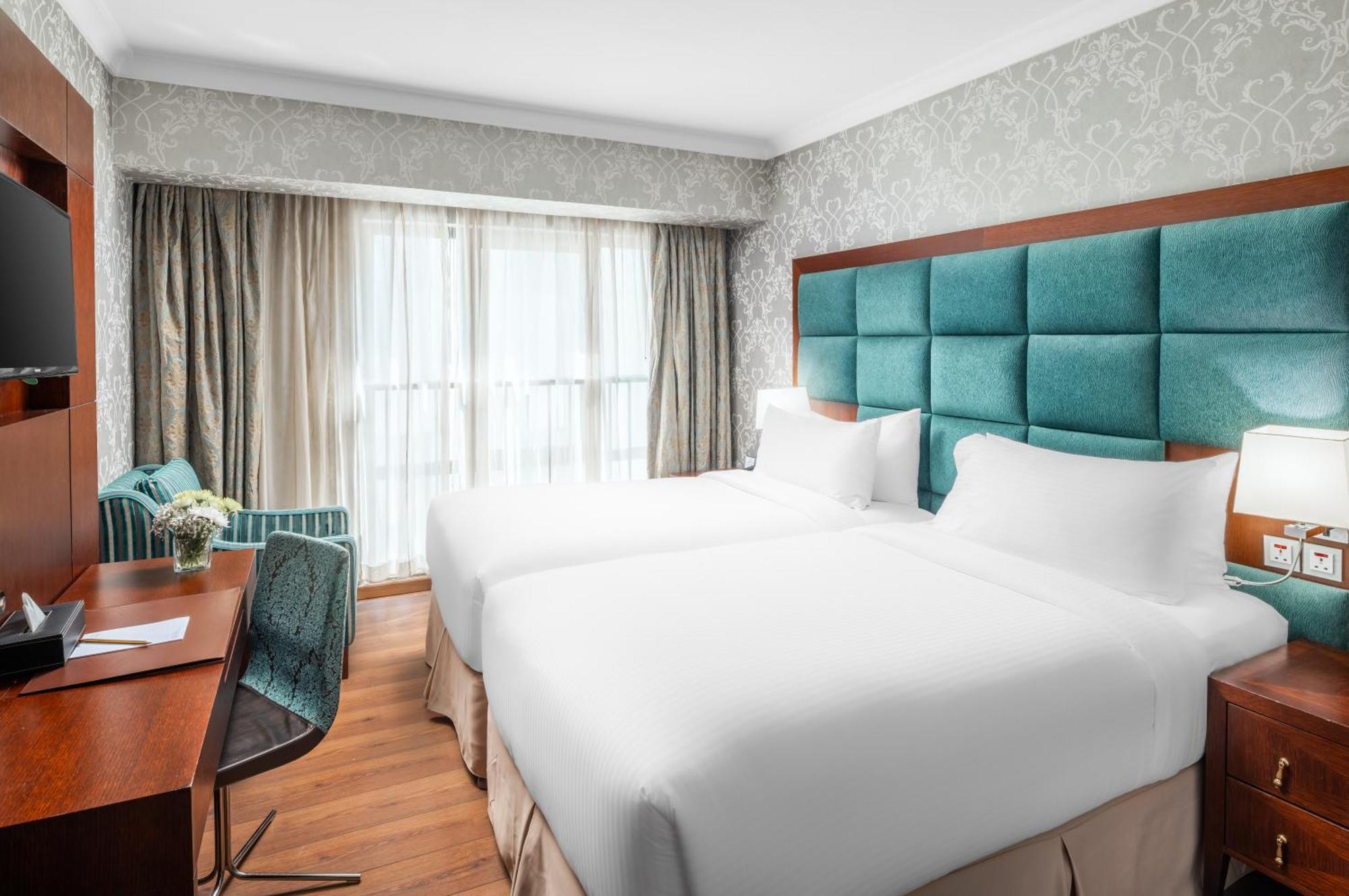 Al Aqeeq Madinah Hotel Медина Екстериор снимка The photo shows a modern hotel room featuring two single beds with white bedding. The headboard is upholstered in a teal fabric. There is a wooden desk and a chair near a window that is covered with sheer curtains and drapes. A cozy armchair in a mat