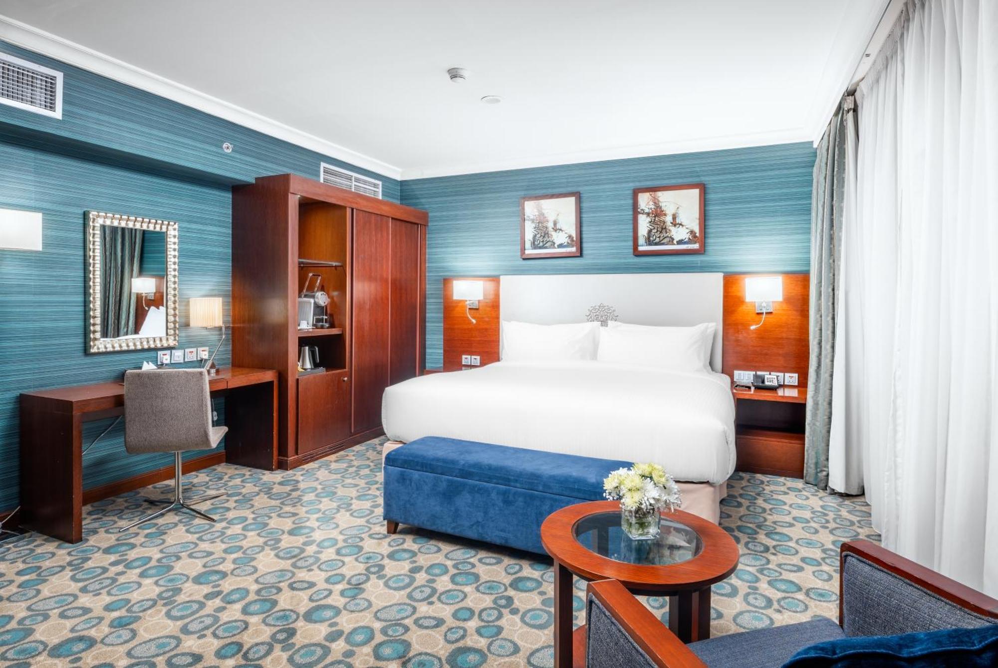 Al Aqeeq Madinah Hotel Медина Екстериор снимка The photo shows a well-decorated hotel room. It features a large bed with white bedding, flanked by wall-mounted lights. Behind the bed, there are two framed pictures hanging on a blue wall. To the left, there's a wooden wardrobe and a desk with a ch