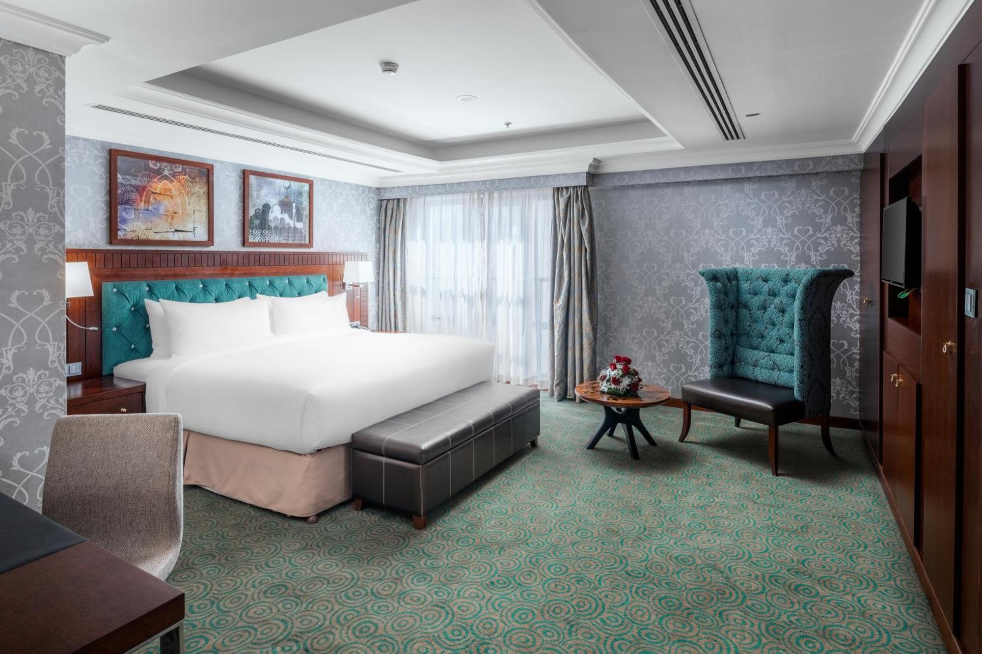 Al Aqeeq Madinah Hotel Медина Екстериор снимка The photo shows a well-appointed hotel room. It features a large bed with a tufted headboard in a turquoise color, complemented by decorative pillows. There is a comfortable ottoman at the foot of the bed and a small coffee table with a vase of flowe