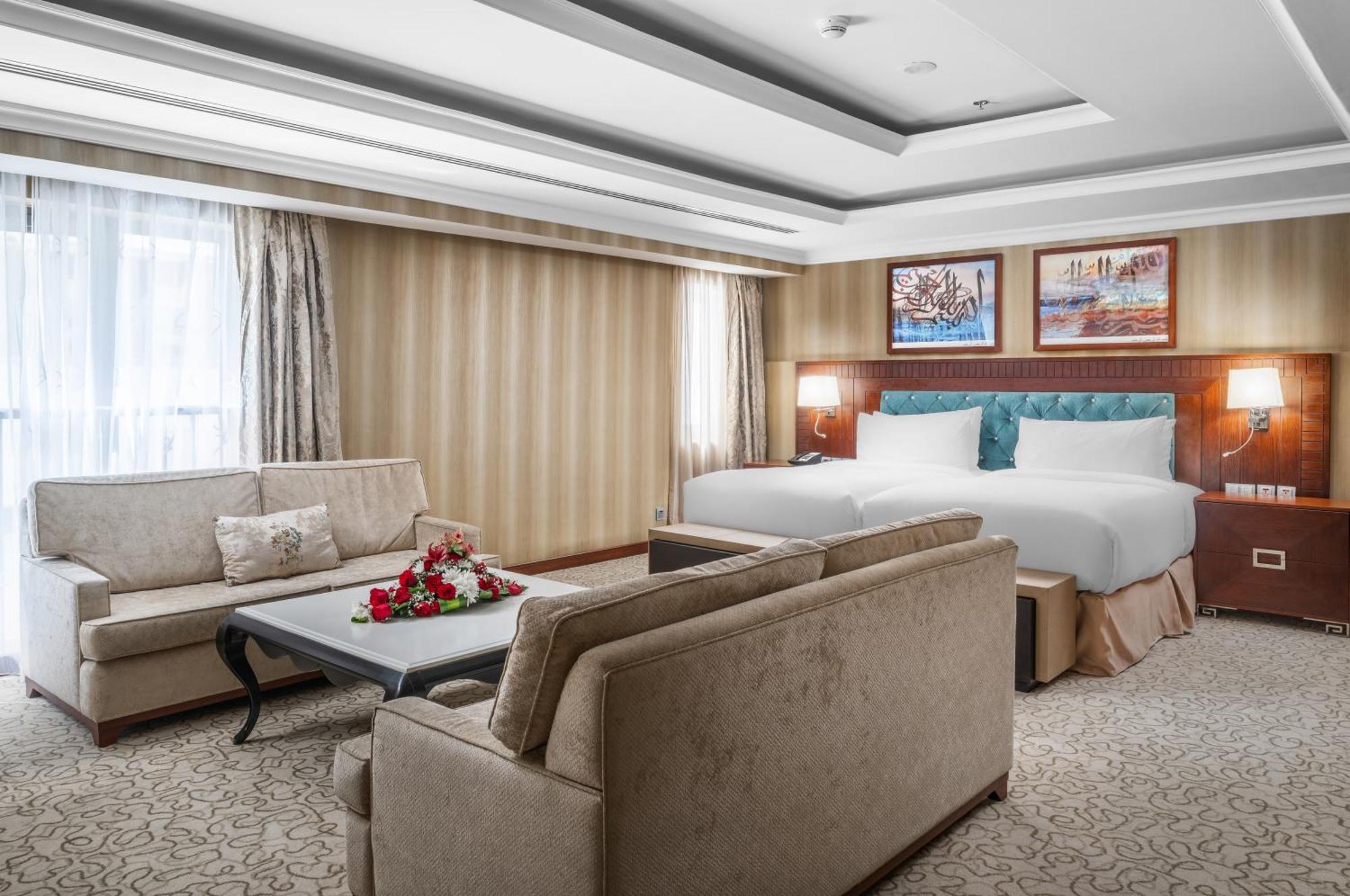 Al Aqeeq Madinah Hotel Медина Екстериор снимка The photo shows a well-decorated hotel room. It features a large bed with two pillows and a light-colored duvet. There are two comfortable couches arranged around a coffee table with a floral centerpiece. The walls have a subtle striped design, and l
