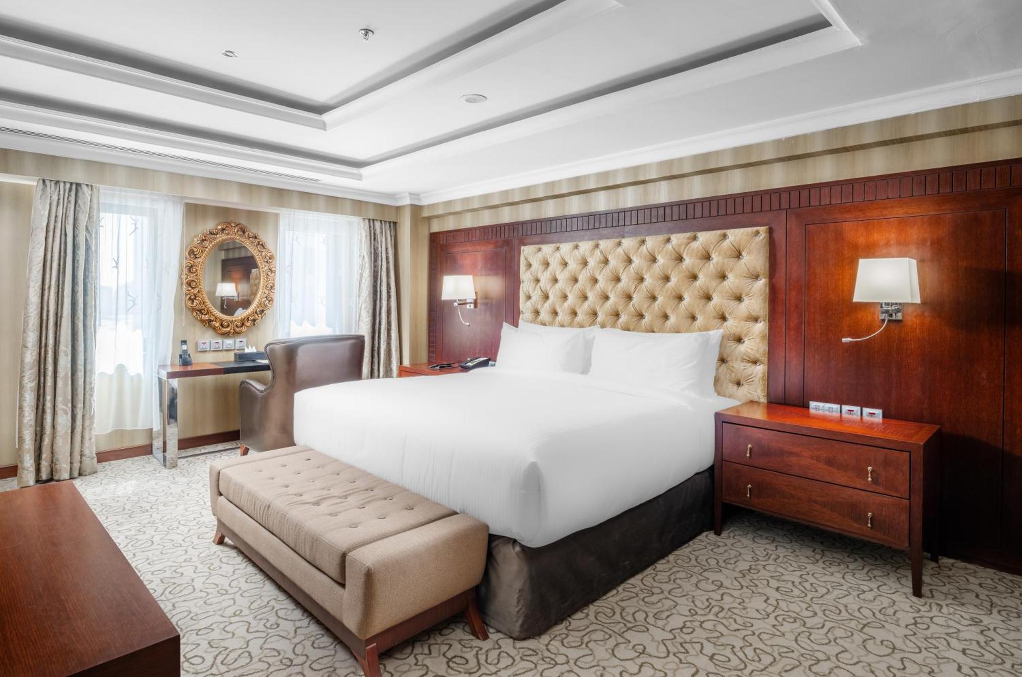 Al Aqeeq Madinah Hotel Медина Екстериор снимка The photo shows a well-furnished hotel room featuring a large bed with a tufted headboard. The room has a warm color palette with wooden accents and soft lighting. There are two windows with curtains that allow natural light to fill the space. Alongs