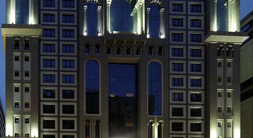 Al Aqeeq Madinah Hotel Медина Екстериор снимка The image shows the facade of a modern high-rise building illuminated at night. The design features large glass windows arranged in a grid pattern, along with decorative architectural elements at the top, including arches and ornate detailing. The co
