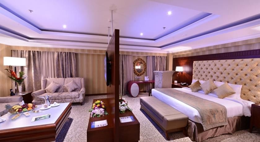 Al Aqeeq Madinah Hotel Медина Екстериор снимка The image shows a luxurious hotel room. It features a large bed with a tufted headboard, flanked by nightstands with lamps. There's a seating area with a plush sofa and a coffee table. A decorative partition separates the bed from the living area. Th