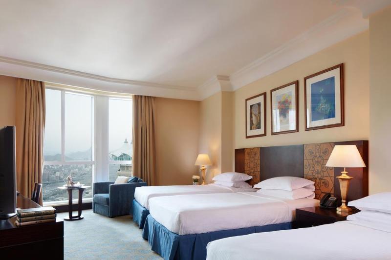 Al Aqeeq Madinah Hotel Медина Екстериор снимка The photo shows a hotel room featuring three beds arranged neatly. The room has a warm, inviting ambiance with beige walls and a soft carpet. Large windows allow natural light to fill the space, offering a view outside. There are curtains in a light 
