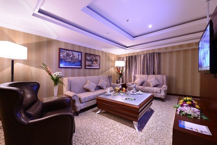 Al Aqeeq Madinah Hotel Медина Екстериор снимка The photo shows a stylish and comfortable living room. It features two light-colored sofas with decorative cushions arranged around a modern coffee table. In one corner, there's a dark, leather armchair. The walls are adorned with framed artwork, and