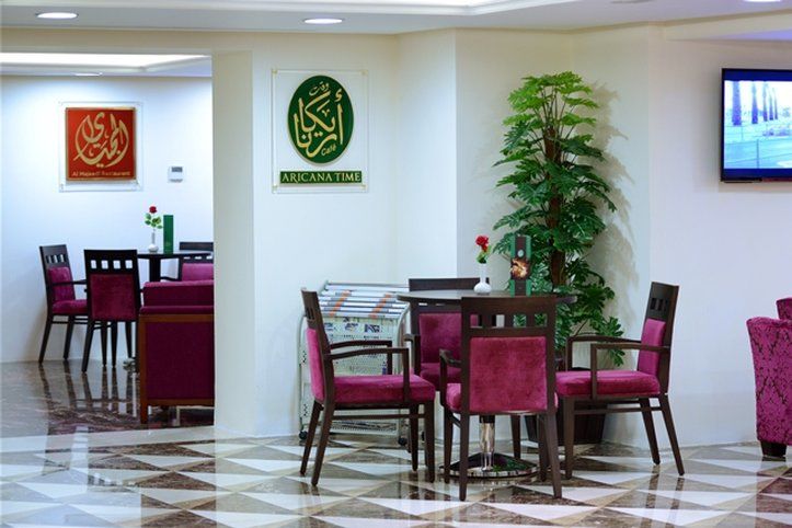 Al Aqeeq Madinah Hotel Медина Екстериор снимка The photo shows an interior space, likely a dining or lounge area. There are several tables with four chairs each, featuring a modern design with dark wood and plush maroon upholstery. On the walls, there are decorative signs, one of which has Arabic