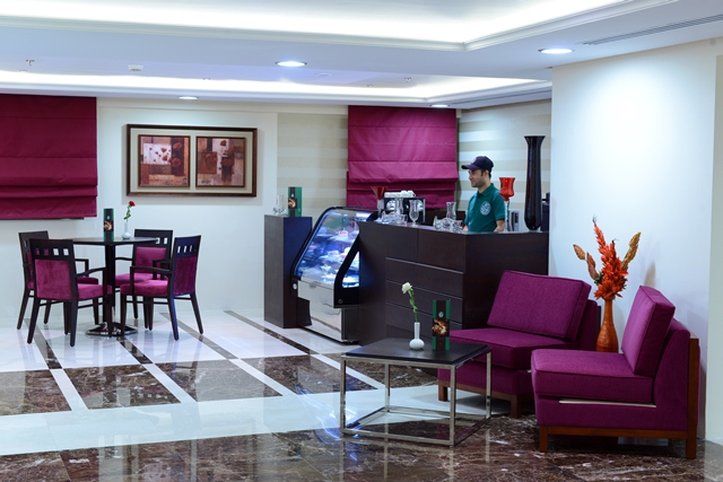 Al Aqeeq Madinah Hotel Медина Екстериор снимка The photo shows a modern interior space, likely a café or small restaurant. It features a seating area with a few tables and chairs, all in a dark wood finish with purple upholstery. There is a counter area with a staff member behind it, possibly ser