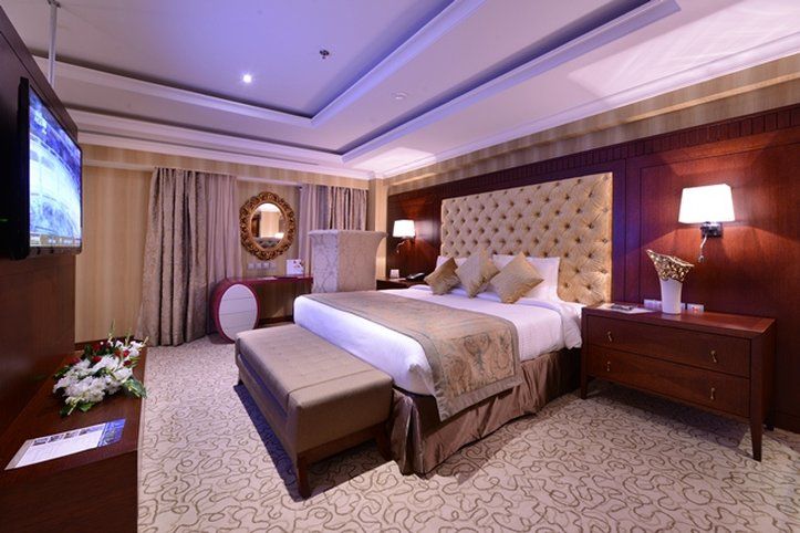 Al Aqeeq Madinah Hotel Медина Екстериор снимка The photo shows an elegantly designed hotel room. It features a large bed with a plush headboard and decorative pillows. On either side of the bed are bedside tables with lamps. There's a comfortable bench at the foot of the bed and a TV mounted on t